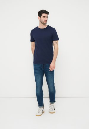 Άνδρας που στέκεται μπροστά σε λευκό φόντο, φοράει μπλε ναυτικό μπλουζάκι, τζιν παντελόνια που είναι σηκωμένα και λευκά αθλητικά παπούτσια με μαύρες ρίγες.