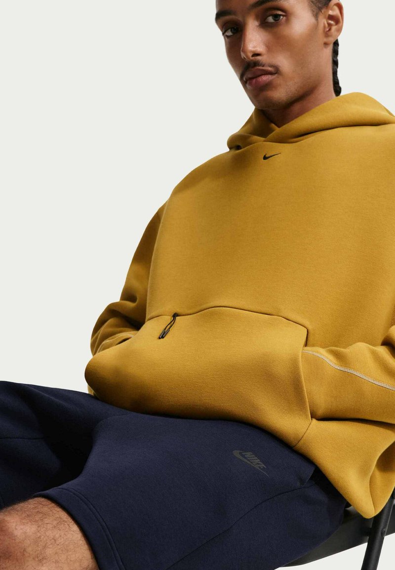 Man met gevlochten haar die een mosterdgele Nike-hoodie en marineblauwe korte broek draagt, zit op een stoel tegen een effen lichte achtergrond.
