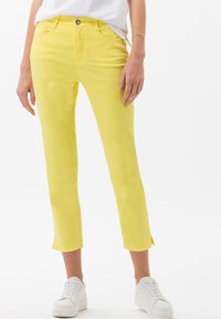 Pantalones cortos amarillos de algodón con ajuste ceñido, que presentan un cierre frontal con botones y aberturas laterales en el dobladillo, combinados con zapatillas blancas.