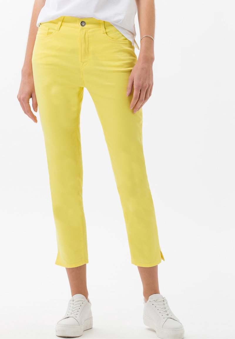 Pantalones cortos amarillos de algodón con ajuste ceñido, que presentan un cierre frontal con botones y aberturas laterales en el dobladillo, combinados con zapatillas blancas.