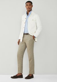 Uomo che indossa una giacca bianca, una camicia a righe blu, pantaloni beige, una cintura marrone e mocassini marrone scuro, in piedi davanti a uno sfondo grigio chiaro.