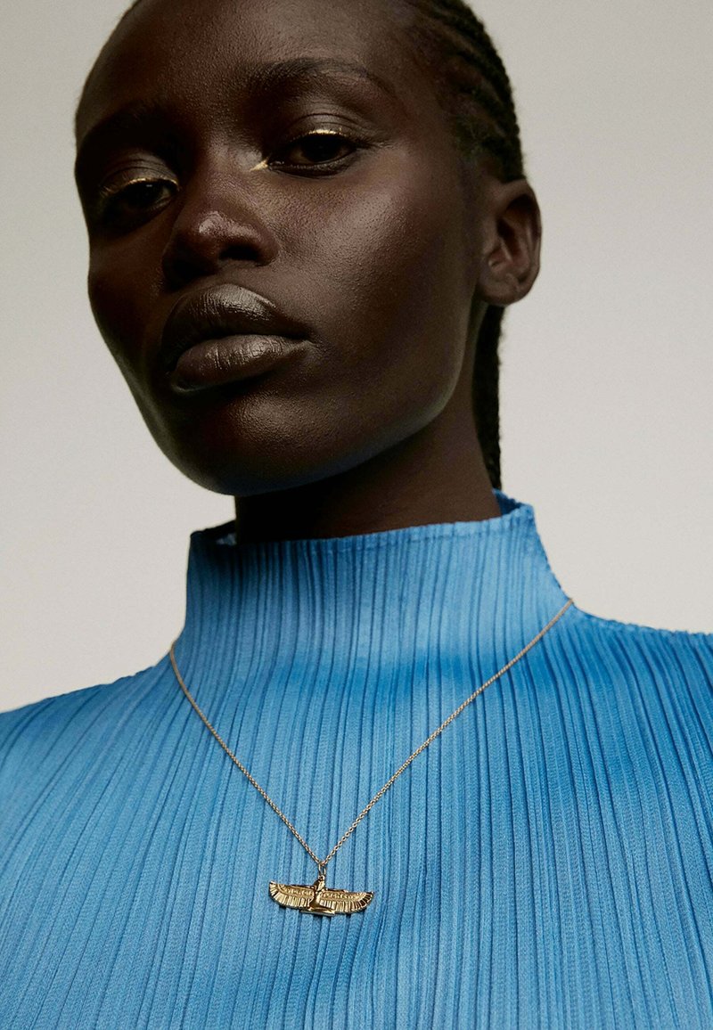 Collar de oro con un colgante alado, lucido sobre una prenda azul plisada de cuello alto, destacando las texturas suaves y los intrincados detalles.