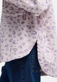 Jasnofioletowa koszula z korduroju z wzorem w panterkę, z zakrzywionym dołem i bocznymi rozcięciami, noszona na ciemnoniebieskich dżinsach.