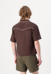 Braunes kurzärmeliges Hemd mit lockerer Passform und weißen Piping-Details an den Schultern, kombiniert mit olivgrünen Shorts.