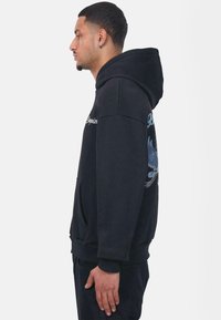Zwarte hoodie met een relaxte pasvorm, met een voorzak en een blauwe vogelafbeelding op de linker mouw. Made van zachte stof.