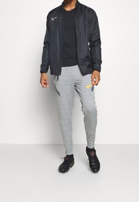 Chaqueta ligera negra, camiseta negra, pantalones de chándal gris con un patrón y acentos en naranja y azul marino, y zapatillas deportivas negras.
