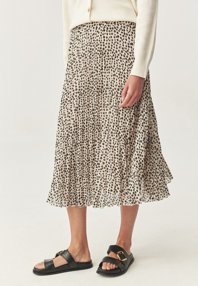Jupe midi plissée beige à pois noirs portée avec un pull crème et des sandales en cuir noir avec des boucles dorées.