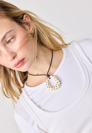 Mlajša ženska s svetlimi lasmi, oblečena v belo majico in z belim ogrlico v obliki kaplje, okrašeno z zlatimi kroglicami na rjavi vrvici.