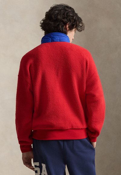 Άνδρας με σκούρα σγουρά μαλλιά φορεί κόκκινο φλις μπουφάν με μπλε γιακά και μπλε παντελόνι με την επιγραφή "USA", με την πλάτη προς την κάμερα.