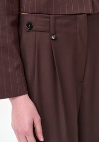 Hand rustend naast bruine op maat gemaakte broek met knoopdetail op het tabblad, gecombineerd met een bijpassende pinstripe blazer mouw.