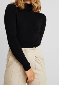 Schwarzer Rollkragenpullover mit langen Ärmeln, aus Strickstoff; getragen mit beigen, hochsitztenden Hosen mit gefältelten Details.