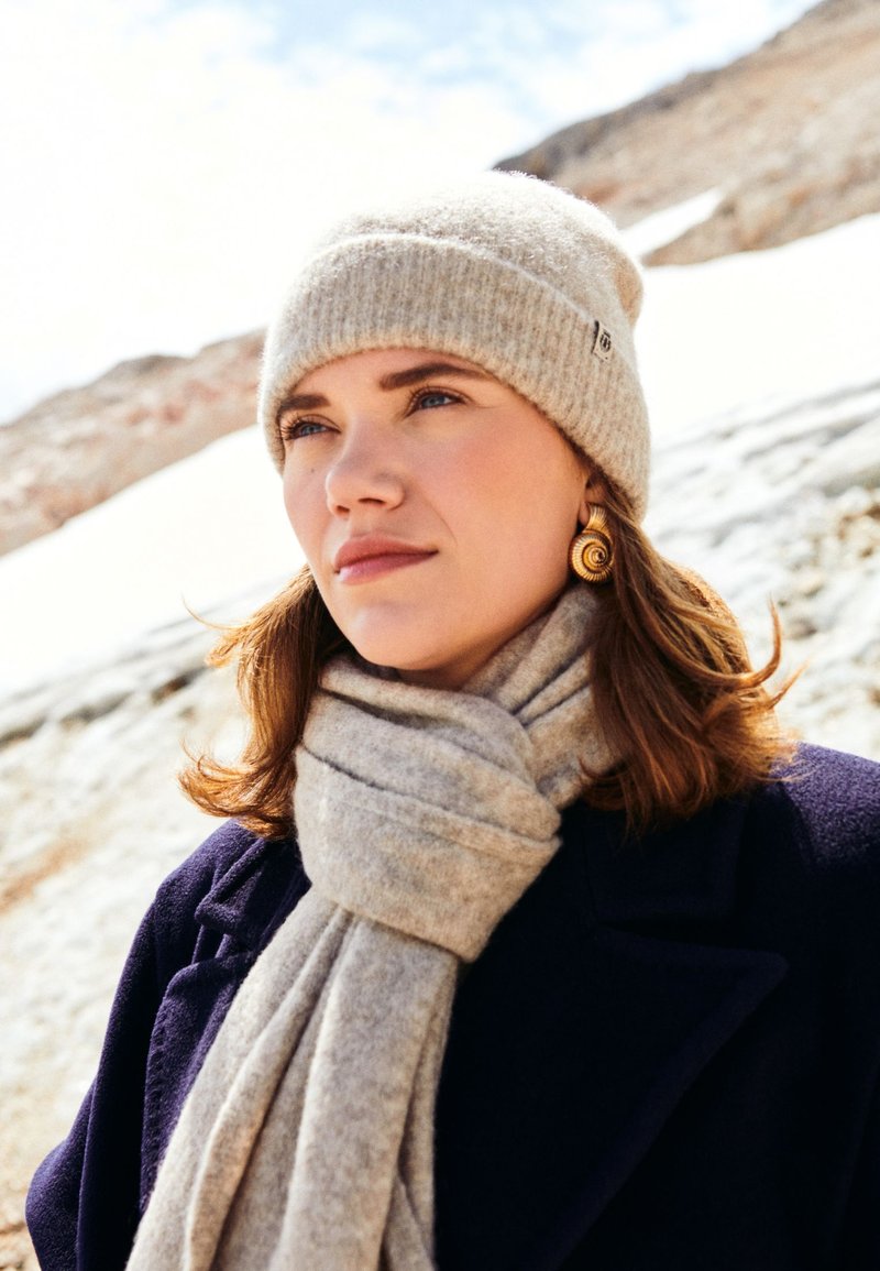 Beige knitted hat, grey scarf, and dark coat. Smooth textures, simple design, and warm materials. Snowy background adds contrast.