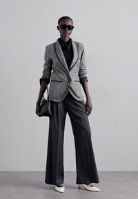 Blazer texturé gris avec bordures noires, chemise noire, pantalon gris à jambes larges et chaussures blanches à bout pointu. Le modèle tient un petit sac à main noir.