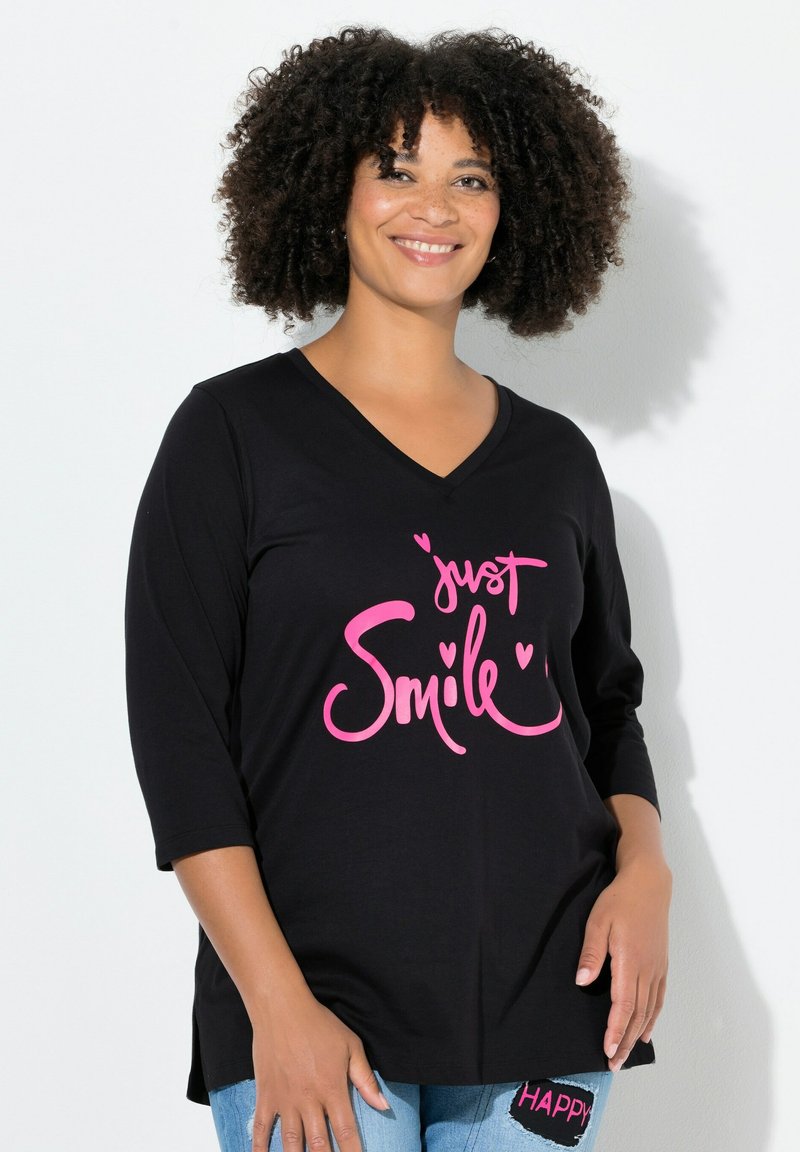 Schwarzes V-Ausschnitt-T-Shirt mit Dreiviertelärmeln, das pinken Text "just Smile" und Herzakzente zeigt. Getragen mit blauen Jeans, die Patch-Details aufweisen.
