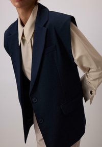 Námornícky bezrukávový blazer s dvomi gombíkmi, s klopenými reverzmi a prednou vreckom, navrstvený na svetlom béžovom tričku so štruktúrovaným golierom.