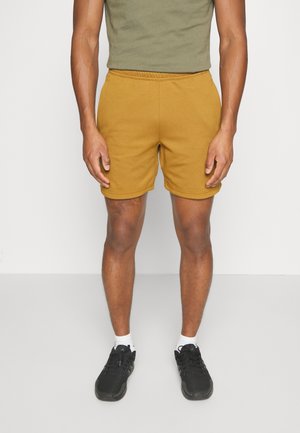 Mężczyzna w musztardowo-żółtych szortach, oliwkowo-zielonej koszulce, białych skarpetkach i czarnych butach sportowych, stojący na jednolitym białym tle.