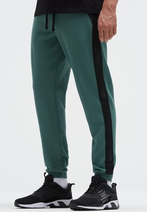 Man wearing teal joggers with black side stripes and black sneakers with white soles, standing on a white background.