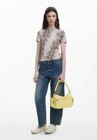 Młoda kobieta ubrana w beżową koszulę w wężowy wzór, luźne niebieskie dżinsy, białe sneakersy, trzymająca żółtą torebkę z wieloma zamkami błyskawicznymi.