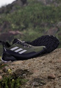 Zapatillas verdes de exterior con detalles en negro, que cuentan con tres rayas blancas, suela texturizada y parte superior transpirable con cordones.