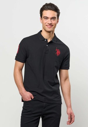 Mlad moški, ki nosi črno polo majico z rdečo vezeno znamko na prsih in rokavu, stoji z eno roko v žepu in se smehlja.