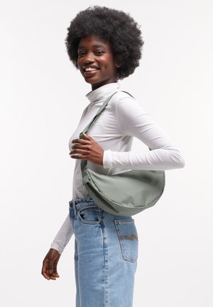 Mujer sonriente con cabello afro que lleva un suéter de cuello alto blanco, jeans azules y un bolso de hombro verde claro.