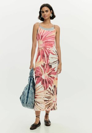 Žena nosiaca bezrukávové midi šaty s kvetinovým vzorom v ružovej a béžovej farbe, drží veľkú modrú rifľovú tašku a nosí čierne šnurovacie topánky.