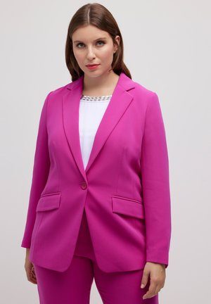 Blazer rose avec un col cranté, fermeture à un bouton et deux poches avant. Porté par-dessus un haut blanc avec un décolleté décoratif.