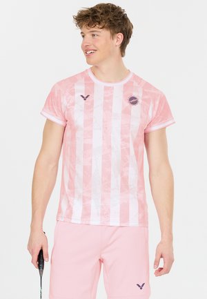 Ung mand iført pink og hvidstribet sportstrøje og pink shorts, der holder en badmintonketcher, kigger til siden og smiler.