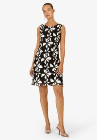 Robe noire sans manches avec broderie florale blanche, présentant une encolure ronde et une coupe légèrement évasée. Texture lisse, design arrivant aux genoux.