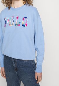 Ljusblå sweatshirt med ordet "POLO" i rutigt tyg med flerfärgad design. Har långa ärmar och en avslappnad passform.