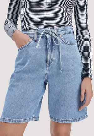 Jeansshorts mit gebundenem Gürtel, getragen zu einem langärmligen schwarz-weiß gestreiften Oberteil, Hand in der Tasche vor neutralem Hintergrund.