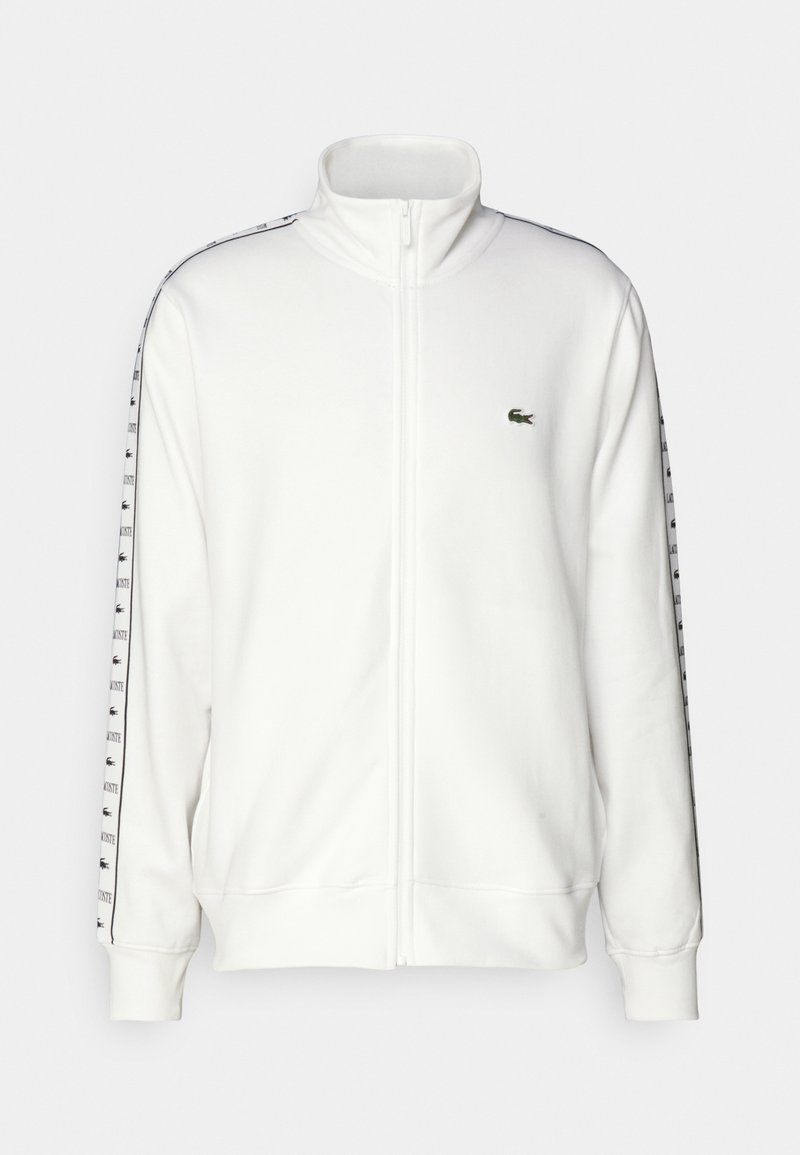 Witte zip-up hoodie gemaakt van zachte stof, met het Lacoste-logo en twee zwarte schouderstrepen met een herhalend tekstpatroon.