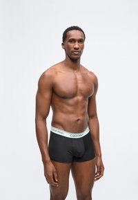 Αθλητικός Μαύρος άνδρας στέκεται, φοράει μαύρα εσώρουχα Calvin Klein boxer με λευκή μέση, ουδέτερη έκφραση, απλό ανοιχτό γκρι φόντο.