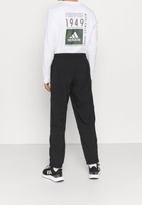 Černé sportovní kalhoty s elastickým pasem, spárované s bílou košilí s dlouhým rukávem, na jejíž zadní straně je velké zelené logo Adidas.