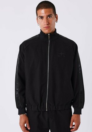 Chaqueta negra con cremallera y cuello alto, puños y dobladillo acanalados. Presenta paneles laterales texturizados y un logo en relieve en el pecho.