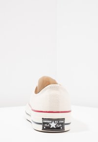 Baskets basses en toile beige avec une semelle en caoutchouc blanc, une bande rouge et un patch logo noir arborant une étoile. Tige texturée avec un renfort au niveau du talon.