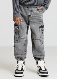 Pantaloni cargo in denim grigio con polsini elastici, tasche laterali e dettagli cuciti, abbinati a sneaker alte bianche e nere.