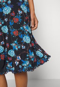 Main tenant le bord d'une robe noire avec des motifs floraux bleus, rouges et blancs et un ourlet à volants.