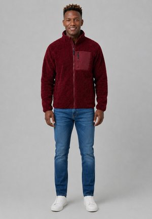 Hombre de pie con chaqueta de forro polar con cremallera color burdeos, vaqueros azules y zapatillas blancas sobre un fondo liso gris claro.
