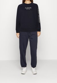 Sudadera azul marino con bordado "Nueva York EST. 1985", combinada con pantalones de chándal a juego. Ambos hechos de una tela suave y texturizada. Zapatillas deportivas blancas.