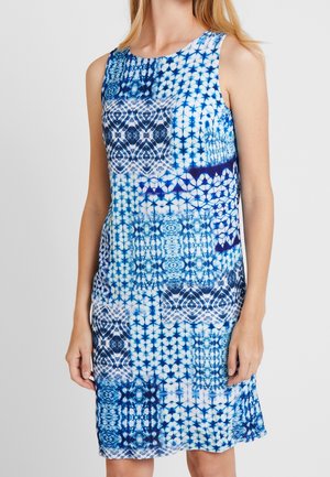Robe sans manches longueur genoux avec des motifs géométriques tie-dye bleus et blancs portée par une femme debout sur un fond uni.