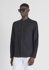 Maglietta nera a maniche lunghe con colletto alla cinese, due tasche sul petto e chiusura a bottoni. Tessuto liscio, indossata con occhiali da sole neri oversize.