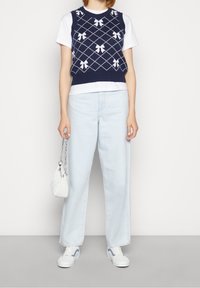 Marineblaues gestricktes Vest mit weißen Schleifenmustern, über einem weißen T-Shirt getragen. Hellblaue weit geschnittene Jeans und weiße Sneakers mit hellblauen Akzenten. Weiße Tasche.