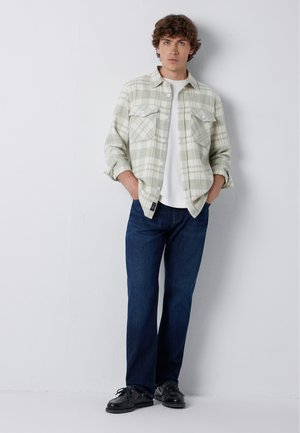 Young man wearing light gray plaid shirt over white t-shirt, blue jeans, and black shoes, standing with hands in pockets against white wall.