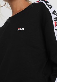 Černá mikina s kulatým výstřihem, na hrudi s malým bílým a červeným logem FILA a kontrastními bílými rukávy s červenými akcenty.