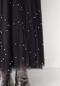Jupe en tulle noire décorée de petites perles argentées portée sur des bottes en cuir noir brillant sur un sol gris.