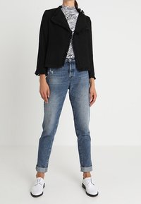 Veste noire texturée avec des bords effilochés, haut à rayures blanches, jeans bleu clair retroussés aux chevilles, et chaussures blanches avec des accents noirs.