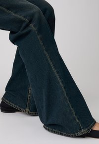 Personne portant un jean denim bleu foncé à jambes larges avec des coutures visibles, associé à des chaussures plates noires sur un fond uni.
