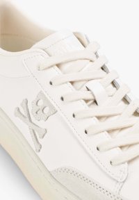 Zapatilla blanca de cuero con cordones crema, detalles en ante y un emblema de calavera y huesos cruzados bordado en el lateral.