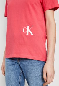 Korálově zbarvené tričko s krátkým rukávem a hladkou texturou, na spodní levé straně má bílé logo "cK". Nošeno s modrými džínami.